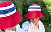 Bildet viser en heklet bøttehatt i rødt, hvitt og blått, brukt som sommerlig supporterhatt til fotball-VM. Bøttehatten har fargene fra det norske flagget, med rød topp og brem, hvite striper og et blått felt rundt midten.