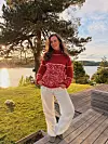 En kvinne står utendørs i kveldssol ved vannet, iført en rød strikkegenser med lyst bladmønster og lyse bukser. I bakgrunnen er det furutrær, plen, terrasse og utsikt over fjorden.