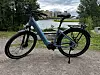 Blå Momas-elsykkel parkert på grusvei ved vannet, med trær og bygninger bak.