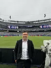 Mats Arntzen i mørk dress står foran banen i et stort fotballstadion med fulle tribuner.
