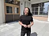 En mann i svart t-skjorte og bukse står på en solfylt gårdsplass foran en bygning.