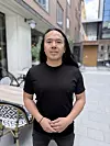En mann i svart T-skjorte står foran moderne bygninger i en gate med uteservering.