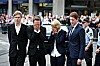 Fire personer i mørke klær står samlet foran en folkemengde utenfor en bygning.