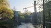 To byggekraner og et høyt industribygg ved en elv med trær i solnedgang.
