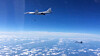 To militære fly i luften over hvite skyer under en blå himmel.