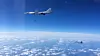 To militære fly i luften over hvite skyer under en blå himmel.