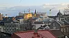 Byutsikt med tak, kraner og kirketårn i bakgrunnen under en overskyet himmel.
