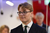 Ung mann i mørk dress, hvit skjorte og slips står innendørs med uskarpe personer i bakgrunnen.