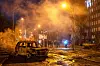 Utbrent bil i en gate med røyk, lys og mennesker i bakgrunnen i Kyiv.
