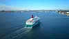 Et stort cruiseskip seiler på fjorden med kjølvann etter seg og byen i horisonten.