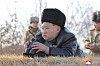 Kim Jong Un ligger på bakken og ser gjennom kikkert mens soldater står i bakgrunnen utendørs.