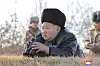 Kim Jong Un ligger på bakken og ser gjennom kikkert mens soldater står i bakgrunnen utendørs.
