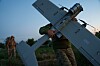 En soldat bærer en stor grå drone på et jorde i skumringen.