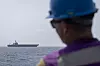 Sjømann med hjelm ser et hangarskip langt ute på havet fra et krigsskip.