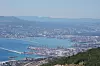 Utsikt over en havn med kraner, båter og en kystby i bakgrunnen.
