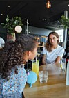 To kvinner sitter ved et trebord på en kafé med drikkevarer og et lyst, moderne interiør i bakgrunnen.