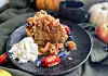 Et stykke Dumlefylt eplekake med crumble serveres på en mørk tallerken med pisket krem, eplebiter og karamellaktig Dumlefyll. Rundt kaken ligger Dumle-biter, epler og høstlige elementer som understreker smaken av eplekake med karamell og kanel.