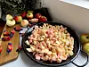 Oppskårne epler ligger klare i en stor panne, omgitt av hele og skrelte epler samt Dumle-biter. Bildet viser forberedelsene til Dumlefylt eplekake, der eplene skal varmes sammen med Dumle og smør til et mykt fyll.