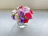 En liten bukett med rosa, lilla og røde blomster i en gjennomsiktig glassvase på et bord.