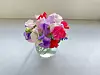 En liten bukett med rosa, lilla og røde blomster i en gjennomsiktig glassvase på et bord.
