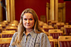 Frøya Sjursæther står i en tom stortingssal med røde stoler og trepanel.
