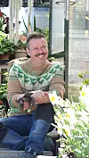 En mann sitter på en benk i et drivhus omgitt av blomster og grønne planter.