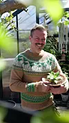 En smilende mann holder en potte med en liten grønn plante i et drivhus.