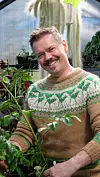 Mann smiler i en strikkegenser med grønt plante- og blomsterinspirert mønster blant potteplanter.
