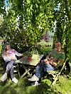 To personer sitter på benker ved et grønt bord under et tett bladverk i en hage.