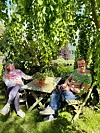 To personer sitter i hagestoler ved et bord under et tett grønt løvverk.