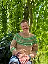 En smilende mann sitter under hengende grønne blader og har på seg en beige genser med grønt mønster.
