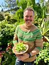 En smilende mann står i en frodig hage og holder en liten plante i en skål.