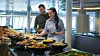 Personer ved et buffetbord med skåler med varm mat og salater i et lyst innendørs serveringsområde.
