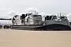 Et stort LCAC-hovercraft står på stranden mens flere uniformerte personell ser på ved en militærbase.