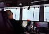 Kontrollrom med operatør foran skjermer og instrumenter, med utsikt mot vann og kaianlegg.