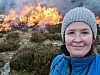 Kvinne i blå jakke står foran en lyngbrann med flammer og røyk i bakgrunnen.
