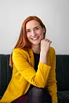 Smilende kvinne med rødt hår i gul blazer sitter på en mørk sofa foran en lys vegg.