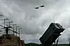Et Patriot-luftvernsystem med radar og missilramper under en mørk himmel, med to fly i luften.