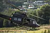 Mørk Bell 412-helikopter i lav høyde over gressfelt med hus og skog bak.