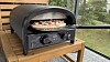 Svart pizzaovn på et trebord med pizza inne i ovnen, plassert på en terrasse.