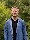 En smilende mann står utendørs foran en grønn, uskarp bakgrunn.