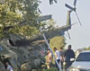 Et militært helikopter ligger skjevt ved en vei mens flere personer står ved siden av en bil.