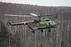 Grønt militærhelikopter med rotor i lufta over en skog.