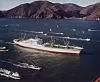 NS Savannah seiler i åpent farvann med mindre båter rundt seg og fjell i bakgrunnen.