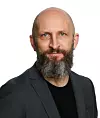 Mann med skjegg og mørk dress poserer mot hvit bakgrunn.