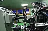 Robotic arm and lab equipment in an indoor workspace with a person standing in the background.