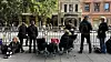 Personer sitter og står ved utendørsbord langs en gate med bygninger og trær i bakgrunnen.