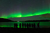 Grønt nordlys strekker seg over himmelen over en mørk fjord, med mennesker i silhuett nederst.