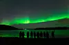 Grønt nordlys strekker seg over himmelen over en mørk fjord, med mennesker i silhuett nederst.