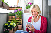 Eldre kvinne i rød cardigan bruker smarttelefon på en balkong omgitt av grønne planter.